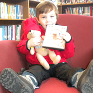 a toddler is holding a teddy bear and book while sitting on a big red chair in the library