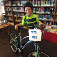 Inside the library is a smiling child wearing bicycle helmet and standing behind a bicycle with a 
